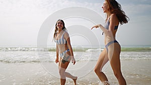 Tanned young women walking, laughing at sea water waves. Two slim girls having fun at seaside. Pretty models in swimwear