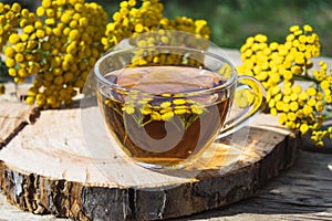 Tansy infusion in a glass cup and yellow tansy flowers on a wooden table. Tansy Herbal tea. Healing herbs