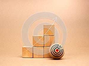 Target icon on wood ball near wooden cube blocks bar graph chart steps with rise up arrows on beige background. Business growth