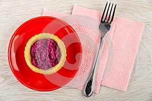 Tartlet with beetroot salad in saucer, fork on paper napkin on wooden table. Top view
