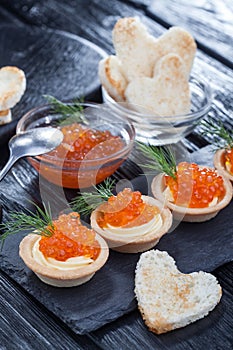 Tartlets with butter and red caviar and dill on a black wooden table.