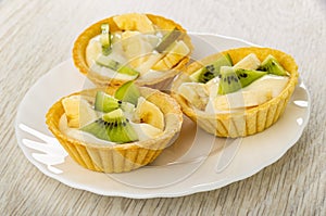 Tartlets with slices of kiwi, banana and yogurt in white plate on wooden table