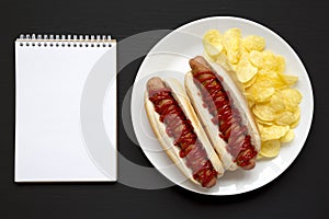 Tasty American Hot Dog with Potato Chips on a white plate on a black surface, top view. Flat lay, overhead, from above