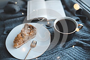 Tasty apple pie on white plate with cup of black tea and open paper book on knit woolen cloth fabric closeup over Christmas glow l