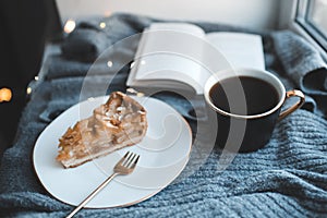 Tasty apple pie on white plate with cup of black tea and open paper book on knit woolen cloth fabric closeup over Christmas glow l