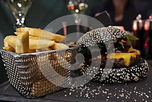 Tasty black burger and french fries in basket served on black cutting board. Selective focus. Copy-space flag stuck into bun