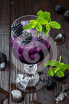 Tasty blackberry cocktail in wine glass with mint and ice on dark wooden table. Summer berry lemonade