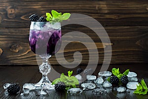 Tasty blackberry cocktail in wine glass with mint and ice on dark wooden table. Summer berry lemonade