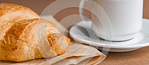 Tasty breackfast. French croissant served on craft paper and cup of black coffee or espresso on brown background. Banner