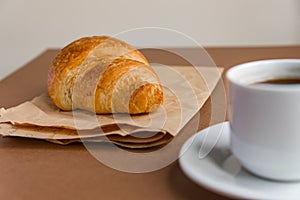 Tasty breackfast. French croissant served on craft paper and cup of black coffee or espresso on brown background. Copy space