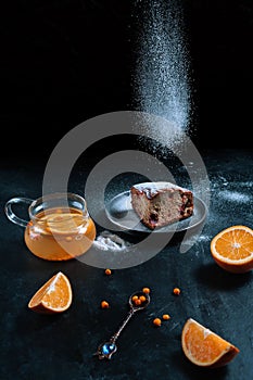 Tasty cake with flying icing sugar and a teapot of sea buckthorn tea with oranges on a dark background.