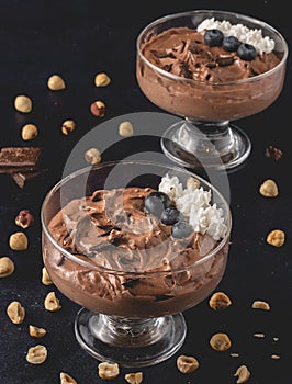 Tasty chocolate mousse dessert placed on dark background with hazelnuts, dark chocolate, blueberries and whipped cream decoration.