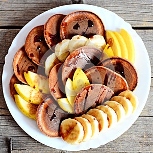 Tasty chocolate pancakes with fresh fruit. Baked chocolate pancakes with syrup, sliced bananas and apples on a white plate