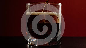 Tasty cola with bubbles is poured into a short glass with ice cubes on a black table against dark red background