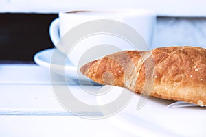 Tasty croissant on a white plate and a cup of coffee on a wooden bench