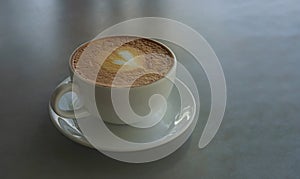 Tasty drinking, a cup of capuccino coffee decorated with heart pattern on brown milk froth in white ceramic cup on gray table