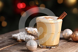 Tasty eggnog with cinnamon, cookies and baubles on wooden table against blurred festive lights. Space for text