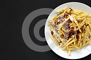 Tasty fast food: french fries with cheese sauce and bacon on a white plate over black background. Flat lay, from above, overhead.