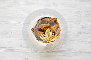 Tasty fast food: fried chicken legs, spicy wings, French fries and chicken fingers in paper box over white wooden background, top