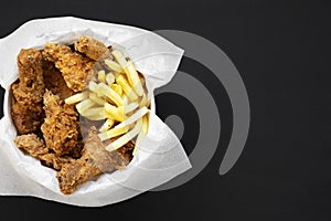 Tasty fastfood: fried chicken drumsticks, spicy wings, French fries and chicken fingers in paper box over black background, top vi