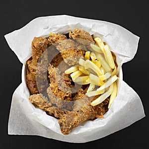 Tasty fastfood: fried chicken legs, spicy wings, French fries and chicken fingers in paper box over dark black background, top