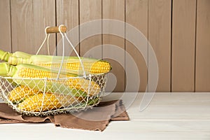 Tasty fresh corn cobs in metal basket on white wooden table, space for text