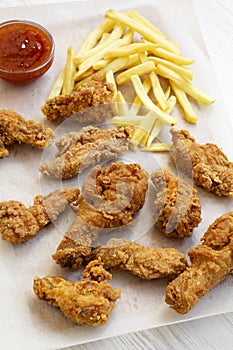 Tasty fried chicken drumsticks, spicy wings, French fries and chicken fingers with sour-sweet sauce on baking sheet over white woo