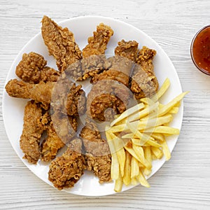 Tasty fried chicken drumsticks, spicy wings, French fries, chicken tenders and sauce on white plate over white wooden surface, top