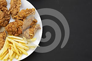 Tasty fried chicken drumsticks, spicy wings, French fries, chicken fingers on white plate over black surface, top view. Flat lay,
