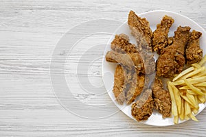 Tasty fried chicken legs, spicy wings, French fries and chicken fingers on white plate over white wooden background, top view.