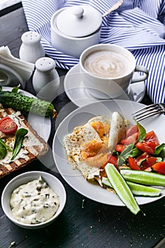 Tasty and healthy breakfast. Fried eggs, vegetable salad, toasts with cream cheese and arugula and cappuccino on a dark background