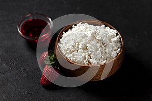 Tasty homemade cottage cheese in a wooden plate with strawberries and jam on a dark background, healthy breakfast on a black table