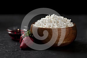 Tasty homemade cottage cheese in a wooden plate with strawberries and jam on a dark background, healthy breakfast on a black table