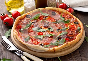 Tasty homemade pizza with ham, tomatoes, cheese, olives and herbs on wooden table.