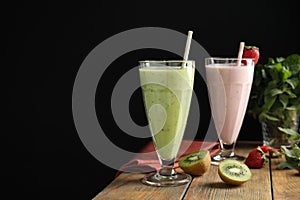 Tasty kiwi and strawberry milk shakes on wooden table against black background, space for text