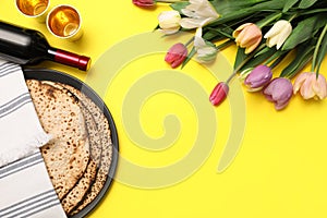 Tasty matzos, wine and fresh flowers on yellow background, space for text. Passover Pesach celebration