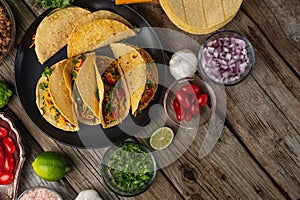 Tasty mexican tacos on the black round plate on the wooden background. Tomatoes, oil, spices, lime are on the table. Traditional