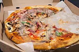 Tasty pizza in a box on a black background. Close-up. Pizza with ham, meat, cheese, tomatoes and crab. There are no two pieces.