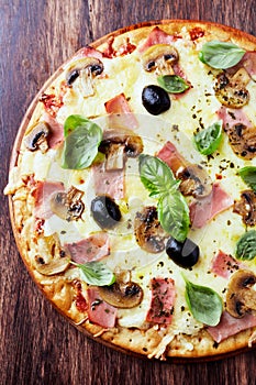 Tasty pizza with ham, champignon mushrooms, mozzarella cheese, black olives and fresh basil on wooden background.