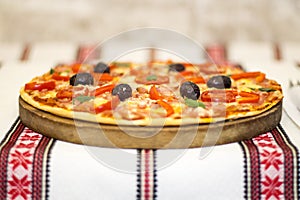 Tasty pizza with vegetables, basil, olives, tomatoes, green pepper on cutting board, table cloth traditional colorful