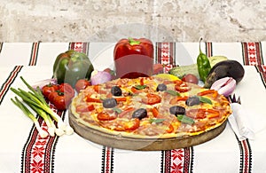 Tasty pizza with vegetables, basil, olives, tomatoes, green pepper on cutting board, table cloth traditional colorful