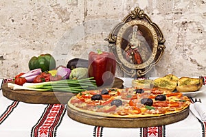 Tasty pizza with vegetables, basil, olives, tomatoes, green pepper on cutting board, table cloth traditional colorful