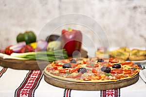 Tasty pizza with vegetables, basil, olives, tomatoes, green pepper on cutting board, table cloth traditional colorful