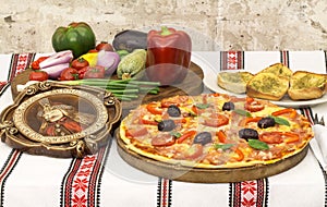 Tasty pizza with vegetables, basil, olives, tomatoes, green pepper on cutting board, table cloth traditional colorful