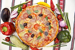 Tasty pizza with vegetables, basil, olives, tomatoes, green pepper on cutting board, table cloth traditional colorful