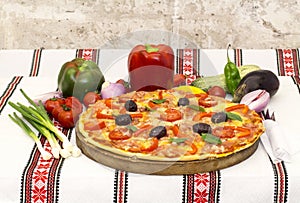 Tasty pizza with vegetables, basil, olives, tomatoes, green pepper on cutting board, table cloth traditional colorful