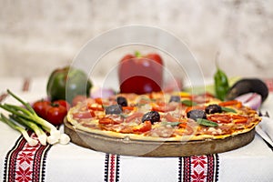 Tasty pizza with vegetables, basil, olives, tomatoes, green pepper on cutting board, table cloth traditional colorful