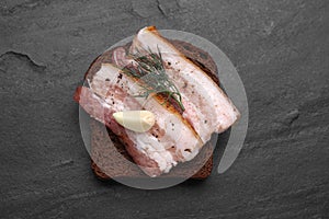 Tasty pork fatback with rye bread slice, garlic and dill on black table, top view