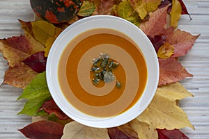 Tasty pumpkin bright orange soup with green seeds served on white plate and autumnal colorful leaves