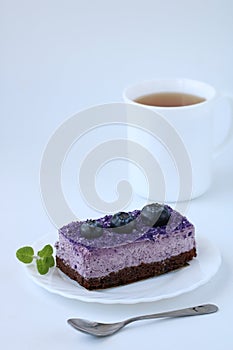 Tasty purple blueberry mousse cake with blueberry on white plate with teaspoon and cup of tea white background, vertical